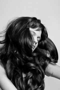 Black and white portrait of a person with voluminous, wavy hair covering their face, posing with a hand on their cheek.