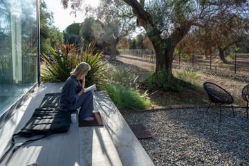 Woman in casual attire enjoying a book on a sunny patio with natural landscaping.