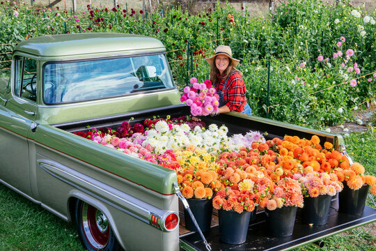A woman smiles behind a vintage truck loaded with vibrant flowers in a lush garden.