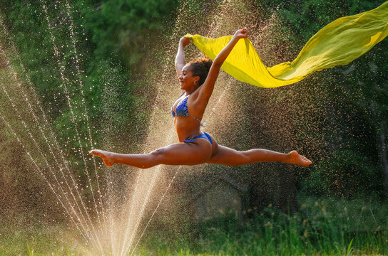 Energetic young woman performs a split jump holding a yellow fabric against a backdrop of water droplets and greenery.