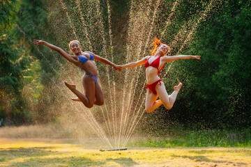 Two joyful women jumping over a sprinkler on a sunny day, with water droplets suspended in the air around them.