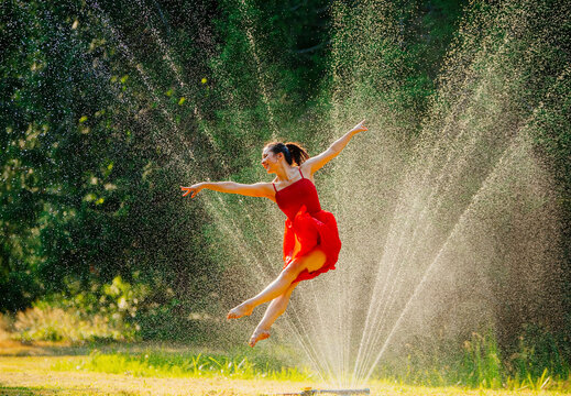 A ballerina in a red dress performs a graceful jump amidst a spray of water with sunlight filtering through.