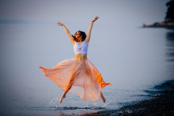 Elegant dancer in an orange skirt performing on a beach at twilight.