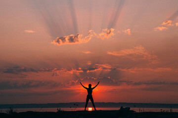 Silhouette of a person with arms raised against a vibrant sunset sky with sunbeams radiating through the clouds.