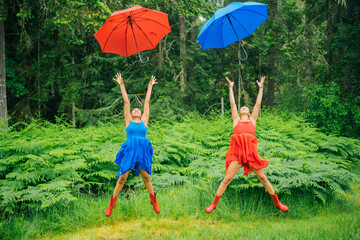 Two joyful women in colorful dresses jumping with red and blue umbrellas against a lush green forest backdrop.