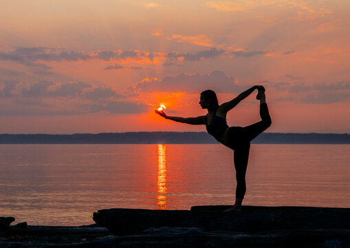 Silhouette of a person practicing yoga on a rocky shore at sunset, holding a balanced pose with the sun in their hand.