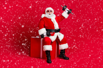 Full length photo of retired old man white beard sit giftbox hold dumbbells shocked easy workout wear x-mas santa costume glove coat belt sunglass cap boot isolated red color background