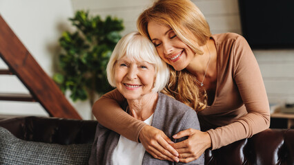 Happy daughter embracing from behind elderly mother at living room