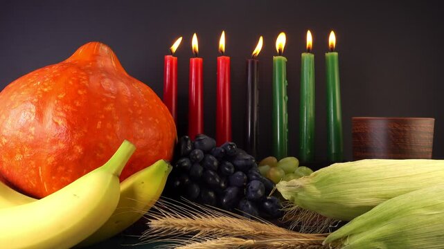 Kwanzaa holiday concept with traditional lit candles, pumpkin, corns, grapes, banana, bowl and wheats over black background, motion
