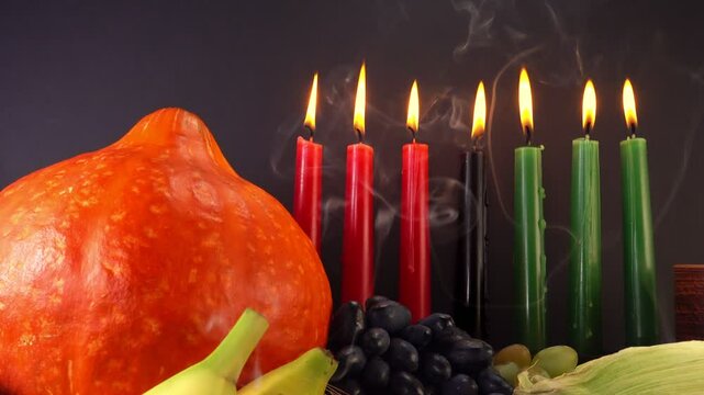 Kwanzaa holiday concept with traditional lit candles, pumpkin, corns, grapes, banana, bowl and wheats over black background and smoke, motion