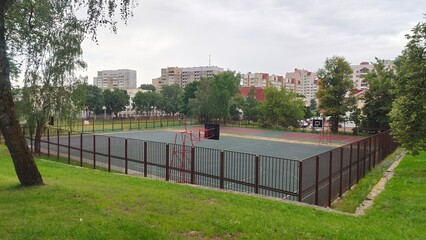 A fenced-in rubberized court has metal structures with basketball backboards and rings with baskets. Nearby are volleyball courts, a grassy lawn, trees and buildings Overcast summer weather and clouds