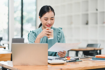 woman in light blue blouse enjoys coffee while reviewing documents at modern office desk. atmosphere is productive and focused, highlighting blend of work and relaxation.