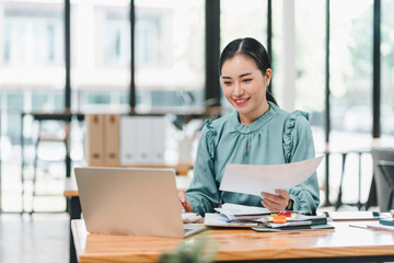 woman is working at desk in modern office, smiling as she reviews documents on her laptop. bright environment enhances her focused and positive demeanor.