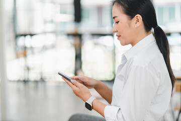 woman in white shirt is sitting and using her smartphone, focused and engaged in her activity. modern setting adds professional touch to her demeanor.