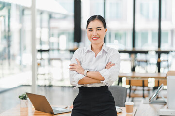 confident businesswoman stands with her arms crossed in modern office environment, showcasing professionalism and approachability. bright workspace features large windows and contemporary furniture,