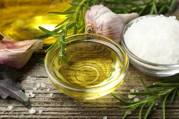 Cooking oil in bowl, salt, garlic and rosemary on wooden table, closeup