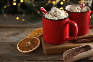 Tasty hot cocoa drink with whipped cream in red mugs and dry orange on wooden table