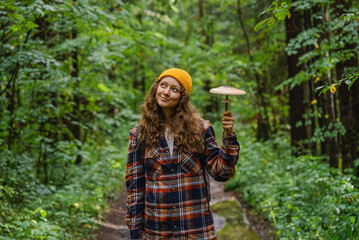 Smiling woman in a yellow beanie and plaid shirt holds a large mushroom while hiking through a lush green forest.