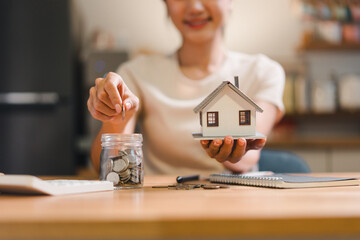 woman is placing coin into jar while holding small model house, symbolizing savings and investment...