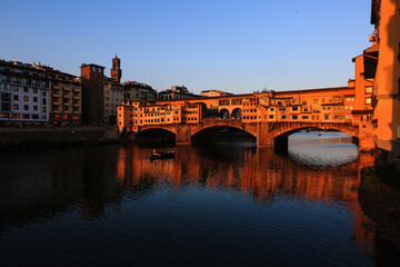 Obraz premium Sunset Over Ponte Vecchio in Florence, Italy The iconic Ponte Vecchio bridge in Florence reflecting the warm sunset glow over the Arno River, with a boat passing underneath.