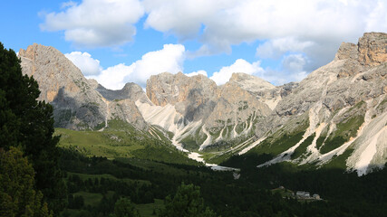 Fototapeta premium Majestic Mountain Peaks in the Dolomites, Italy A stunning view of the rugged mountain peaks and green valleys of the Dolomites in Italy, under a bright blue sky with fluffy clouds.