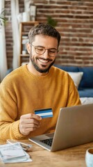 An Indian man with a beard, wearing glasses and a checkered shirt, sits at his desk in a contemporary home office, using a laptop and holding a credit card for online transactions