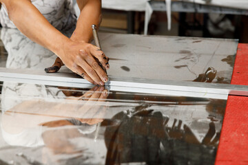 A hand presses down on a glass cutter, tracing a perfect line on a measured sheet of glass inside a...