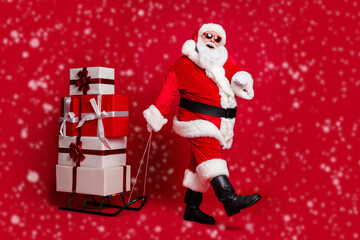 Ho-ho-ho. Full length photo of retired old man white beard hold sled giftbox walk snowy street wear santa x-mas costume coat sunglass headwear black boots isolated red color background