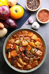 Delicious stew with vegetables in bowl and ingredients on grey table, flat lay