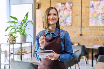 Young woman worker, owner in apron holding notepad pen in restaurant, coffee shop