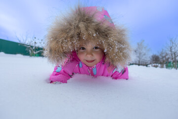 A child in a colorful jumpsuit lies in the snow and looks at the camera, smiling. The child is in the snow, wearing a hood with fluffy fur. The child is crawling in the snow, enjoying the winter.