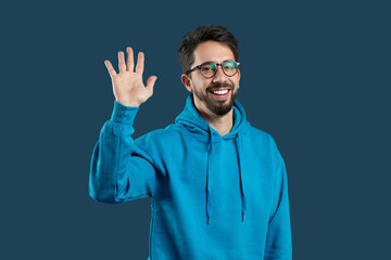 Naklejka premium A cheerful young man greets with a smile and raised hand, dressed in a blue hoodie, set against a dark, solid backdrop. His happy expression conveys friendliness and warmth.