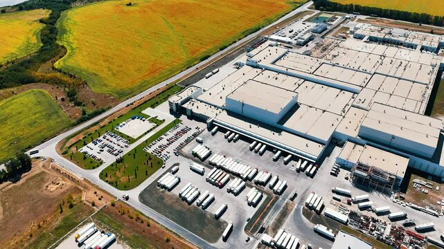 Multiple lorries at the territory of a huge industrial plant. Car park for automobiles is nearby. Aerial perspective on the factory locating in the fields.