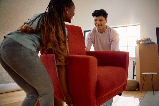 Happy man and his African American girlfriend carrying armchair while relocating into new home.