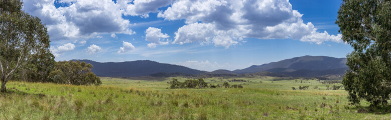 Panorama of Bogong Creek, where peaceful meadows stretch across Namadgi National Park. Lush green grass dotted with boulders and trees, all set against distant hills and a cloudy blue sky.