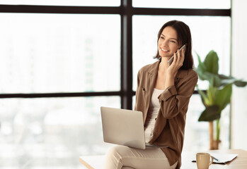 Fototapeta premium Business Communication. Smiling girl making phone call sitting on desk in modern office, using laptop, looking away