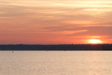 A tranquil sunrise with warm orange and golden hues reflects on the calm water, as a distant shoreline and trees create a serene silhouette.