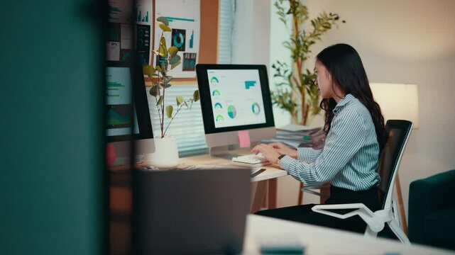 Asian businesswoman sits at her desk, working on a computer. Analyzing data and managing spreadsheets, she demonstrates professionalism in a corporate environment