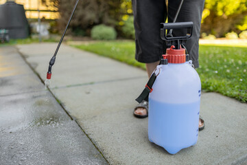 Woman spraying gray concrete patio paving slabs with liquid weed killer from pressurized plastic tank and handheld nozzle, with grass lawn in background
