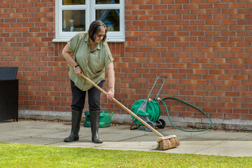 Woman in black trouser pants and wellie boots sweeping concrete slab paved patio after trimming...