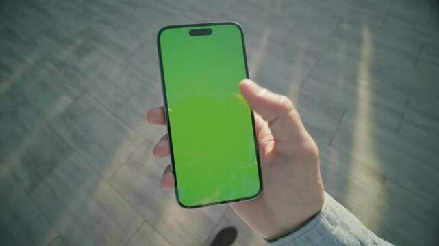 Close up view on hand man holding and scrolling vertically phone with green screen standing. On background sunlight. Typing on smartphone