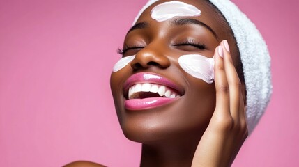 A person with a towel on their head applies a creamy facial product to their skin, beaming with joy in a bright pink setting. Their enthusiasm reflects self-care and wellness