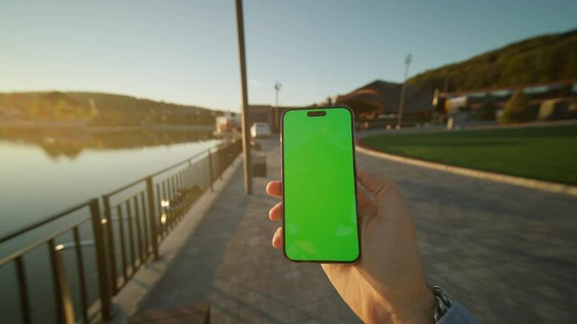View on hands holding vertically phone smartphone device with green screen. On background lake on sunlight. Nature