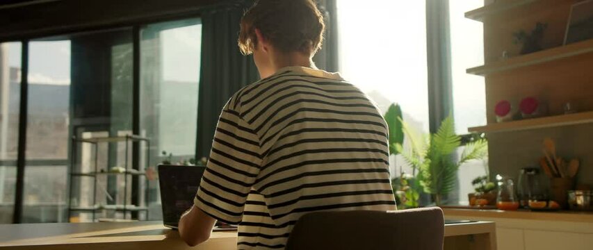 Footage of focused young man looking at display of laptop. Male student working at home. Guy typing on keyboard in kitchen. Moving around tracking shot. Modern interior