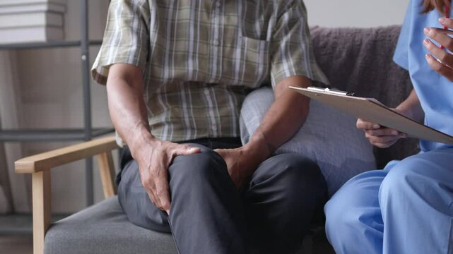 Doctor in blue scrubs is holding a clipboard and taking notes while talking to an elderly male patient about his knee pain