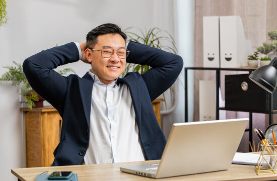 Asian businessman freelancer taking break leaning on chair after working celebrating success victory at modern home office workplace desk. Male Chinese in formal suit puts hands behind head relaxing - Powered by Adobe