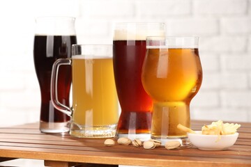 Glasses with different types of beer and snacks on wooden table against white brick wall, closeup