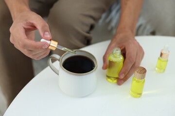 Young man dripping CBD tincture into drink at white table, closeup
