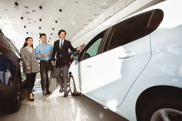 Buying Auto Concept. Successful Car Seller Helping Young Couple Choose New Automobile In Dealership Showroom