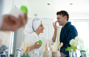 A happy couple in love in bathrobes enjoys a morning skincare routine. The woman applies cream to the man's nose, both smiling in a modern bathroom, reflecting their playful and intimate moment.